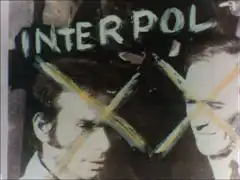 A black and white photograph of two men, their faces crossed out and the word INTERPOL written above them