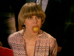 Purdey kneels on the floor with her hands tied behind her back, an apple in her mouth