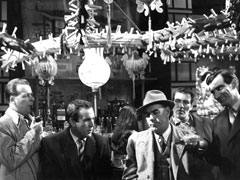Publicity still - The police question Dr. Keel at the Rising Sun: Det Sgt & Dr. Keel on the left, DS Wilson centre with Lila facing away behind him, Bart & Vance, offering Wilson a peanut, on the right