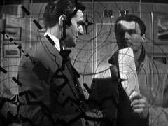 Steed talks to Dr. King, who is gazing at the see-through air controller’s map that is between the camera and them. He taps his chin with some papers