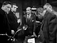 The assembled dealers, standing around the table, look nervous as Fenton conducts his auction of illicit diamonds