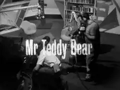 title card: white text reading ‘Mr. Teddy Bear’ superimposed on a wide shot of a TV studio, the crew crouched around the fallen Wayne-Gilley