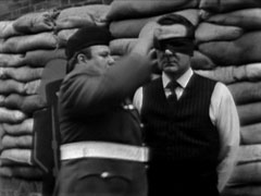 Jessop ensures Steed’s blindfold is straight as he awaits his execution in front of a row of sandbags