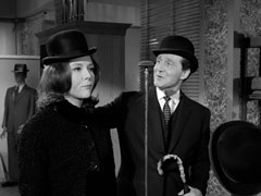 Steed grins as he puts one of the bowler hats on sale in the store on Mrs. Peel’s head