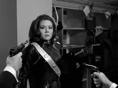 Mrs. Peel backs against a pillar as the villains approach her with their guns pointed at her