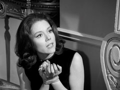 Mrs. Peel leans against the fireplace, her wrists bound with cloth tape