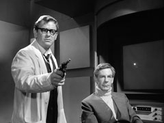 Dr. Armstrong has turned from watching his monitor while Benson stands beside him, pointing his revolver towards the camera - and Steed