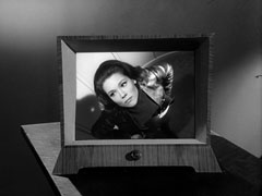 The closed-circuit television monitor shows Mrs. Peel staring up into the camera inside the lift, her hands defiantly on her hips