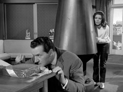 Mrs. Peel takes cover behind the chimney as Steed cautiously opens the box of chocolates