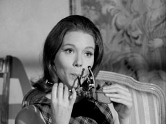 Mrs. Peel blows into a tiny set of bagpipes, behind her an ornate tapestry hangs on the wall