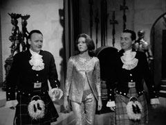 After dinner, Angus leads Mrs. Peel and Steed down an armour and weaponry decorated corridor to see where Black Jamie was walled in. Angus on the left and Steed on the right are in formal Scottish attire with furred sporrans and tight black jackets; Mrs. Peel wears a blue lamé suit without a blouse