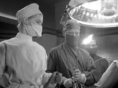 In full surgical garb, Dr. Massie prepares to operate with a blow torch. Miss Thirwell attends on his right while Sager control the gas flow, rather than anaesthetic, in the background. A huge operating theatre light dominates the scene