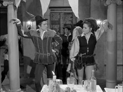 Mrs. Peel leans against a pillar, resplendent in her Robin Hood outfit and critical of Duboys for wearing the same costume; three students in mediaeval costume are huddled behind her on the left