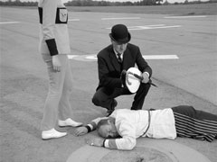 Steed and Mrs. Peel examine the dead milkman lying on the runway