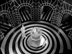 Centre of the maze - Emma stands near a strange, pulsing machine in a circular room with black and white concentric circles on the floor. Around it, strange arched passageways with zigzag and wavy black and whgite designs stretch away in every direction
