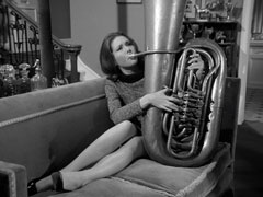 Mrs. Peel lounges on Steed’s sofa, playing his tuba