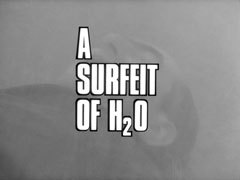 title card: white all caps text reading ‘A SURFEIT OF H20’ outlined in black and superimposed on Barker lying on the ground, gasping for air in the torrent of rain