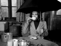 Mrs. Peel sips a cup of tea in Eli’s house while holding an umbrella over her head