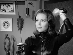 Close-up of Mrs. Peel revealing her face for the first time by removing her fencing mask; she wears a stretch jersey catsuit fronted in vinyl. Her flat, behind her, has an assortment of neo-Classical ornaments