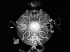 Top-down view of a seance, there are four places around a round table at each of which is a photograph of Steed, the seat at the bottom of the screen is empty