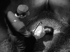 Over the shoulder view of the guard stroking Mrs. Peel’s neck menacingly with his riding crop as she looks up at him from the straw floor