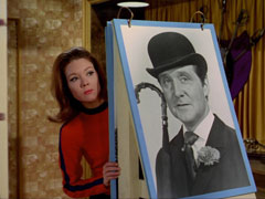Mrs. Peel, in an orange catsuit, peers around the enemy identification chart, currently showing a large black and white photo of Steed