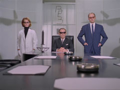 The austere BEB boardroom - all black and white. Pemberton sits at the end of the glossy black table, flanked by Dr. Voss and Gilbert, who stand either side of him; all three wear sunglasses