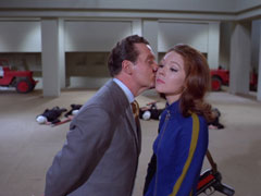 Steed gives Mrs. Peel a light peck on the cheek for saving his life, the dead firing squad she has just mown down with a submachinegun lie on the ground of the underground city behind them