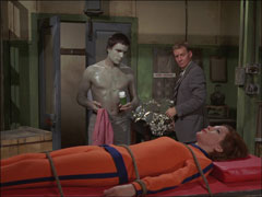 Emma is tied to a table in her orange catsuit, as Dr. Creswell Haworth explain their plot while Creswell sprays Haworth with an insulating compound
