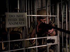 View through the bars of a tiger cage: Nesbitt aiming his rifle to the right of the camera. The cage bears a sign that reads ‘DO NOT FEED THE ANIMALS’