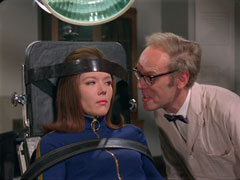 Primble, on the right, gloats at Mrs. Peel who has been strapped to an examining chair by leather belts