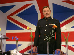 Colonel B stands in his patriotically decorated office, the wall painted as a Union Jack; a row of flowers in vases across his desk represents his field agents