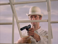 Vickers, dressed in his Classy Glass Cleaning uniform of all white including his bowler hat, points a silenced pistol through the window towards the camera
