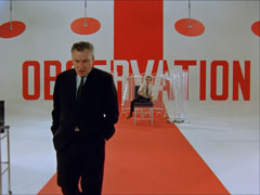 Op art or therapy? Gibbons sits in the middle of a red and white painted room with the word OBSERVATION in huge letters on the far wall, Dr. Reece walks towards us