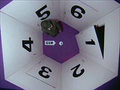 op art or television? A top-down view of Steed standing in a tiny hexagonal room with a purple floor. It has six doors each labelled with the numbers 1–6, clockwise from the right, on the floor is an arrow labelled GUN pointing at a pistol inside a circle in the middle of the room