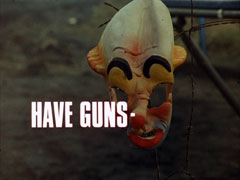 title card: white all caps text reading ‘HAVE GUNS -’ superimposed on a rubber clown mask hanging from a broken barbed wire fence