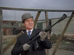 Standing before the fence of the defence installation, Steed holds one of the experimental rifles