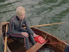 Rhonda sits in the sinking rowing boat in a wetsuit, a red phone on the thwart in front of her