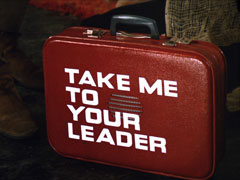 title card: white all caps text reading ‘TAKE ME TO YOUR LEADER’ askew so it’s in line with the red suitcase behind, to make it look like it’s on the side; the suitcase has a speaker in the middle of it