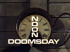 title card: white all caps text reading ‘NOON DOOMSDAY’ - the ‘NOON’ is vertically aligned and horizontally centred - superimposed on a broken railway station clock whose hour hand has been pushed to 12 o’clock by the revolver held in Grant’s gloved hand