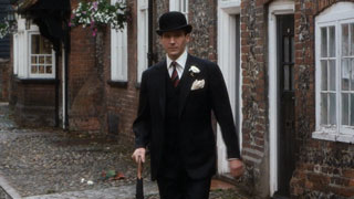 Steed walks down the cobbled pavement of a country village, impeccably dressed in a pinstriped suit and bowler hat