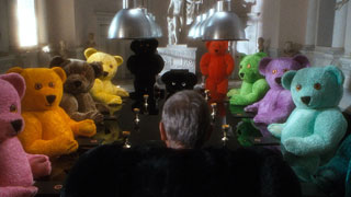 Sir August, with his back to us, addresses a room full of people in different coloured teddy bear costumes
