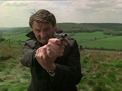Perov stands atop a hill among the fields and steadies his pistol with his other hand around the wrist as he takes aim