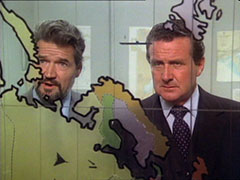 Milroy and Steed stand behind a map painted onto a sheet of glass, much as happened in Dead on Course