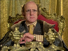 Turner sits on his red and gold throne, a gold curtain behind him; he wears a gold shirt and tie, and wristwatch