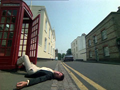 Palmer lies on the pavement, falling out of the door of the red telephone box on the side of the street that stretches away from us, the villains Triumph Dolomite can be seen driving away