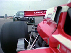 Rear-facing shot on the outside of a March 75B Formula 2 racing car painted in red and white - Steed’s ‘Big Cat’ Jaguar is catching up to it swiftly and is about to pass it