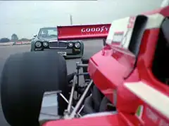 Rear-facing shot on the outside of a March 75B Formula 2 racing car painted in red and white — Steed’s ‘Big Cat’ Jaguar is catching up to it swiftly and is about to pass it