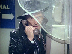 The double agent Murford makes a phone call in the pouring rain, his black hat and overcoat little protection from the elements