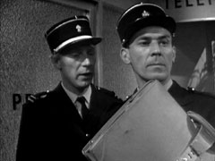 Roland, holding Meyer’s briefcase in the foreground right, is suspicious of Steed when Curly, stading behind him, says Steed had asked to take charge of the briefcase